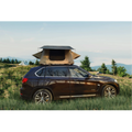 A tan deluxe rooftop tent mounted on a brown SUV parked in a scenic mountain meadow.