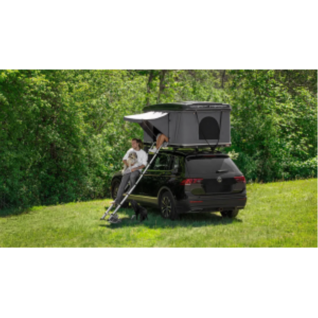 A man and dog sit on a ladder beneath a Room 1916 fiberglass hardshell rooftop tent on a black SUV.