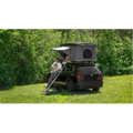 A man and dog sit on a ladder beneath a Room 1916 fiberglass hardshell rooftop tent on a black SUV.