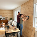A couple smiles as they install the 'Natural' Entrepot La Grange pine wall planks in a sunlit living room renovation.